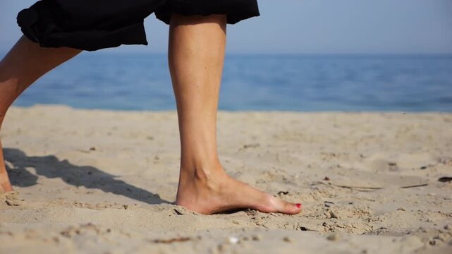 Female feet walking on sand at the beach with seascape at background. Legs of adult woman stepping along sea shore. Barefoot lady strolling near ocean coast. Concept of summer vacation. Low view