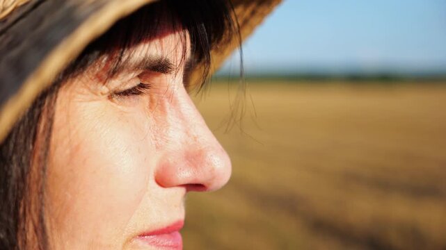 Female farmer in straw hat looking into wheat field and examining plantation at sunset. Close up face of adult beautiful woman illuminating by bright rays of sun. Concept of agriculture business