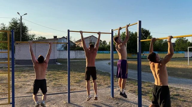 Group of strong sporty men doing pull ups on horizontal bar at sports ground. Young shirtless sportsmen exercising at nature. Muscular athletes training outdoor. Healthy and active lifestyle concept