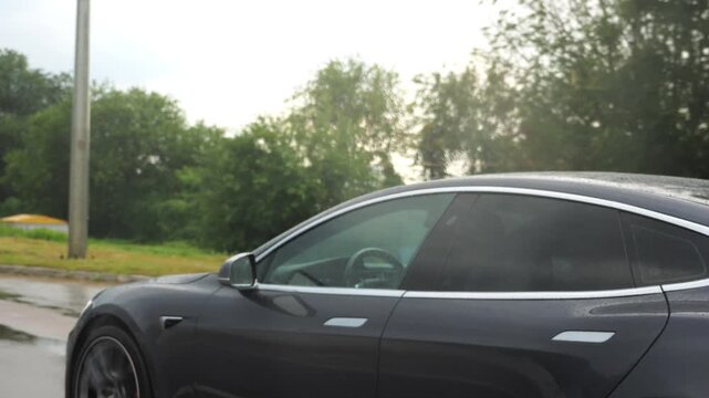 Businessman in shirt driving a modern electric car at sunny rainy day. Male successful entrepreneur riding on electrical vehicle at city. Young manager commuting to work at urban road. Slow motion
