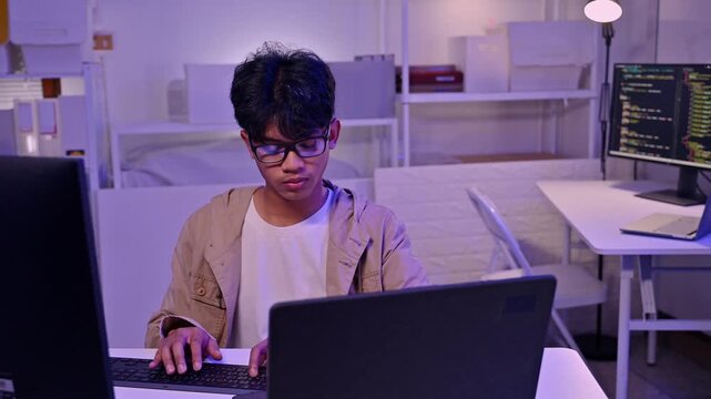 Focused young male programmer wearing glasses working on laptop in modern neon illuminated room