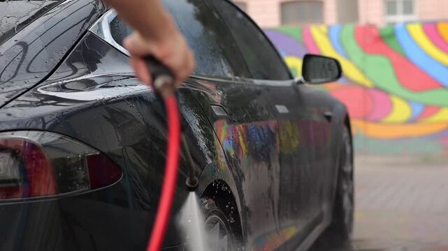 Male hand washing a modern electric auto at a self-service car wash using a high pressure water stream. View captures dynamic splashes highlighting eco friendly transport and urban routine. Slow mo