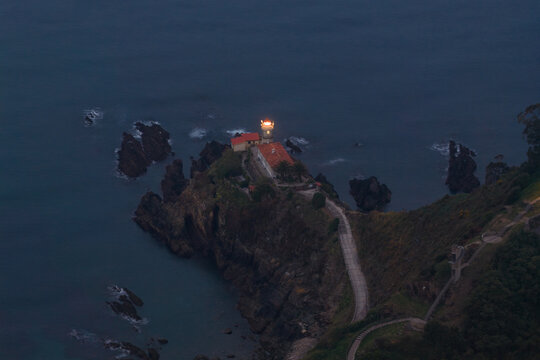 Aerial view of the Cudillero Lighthouse perched on a rugged cliff with its beacon shining over the dark Cantabrian Sea at dusk Cudillero, Asturias, Spain.