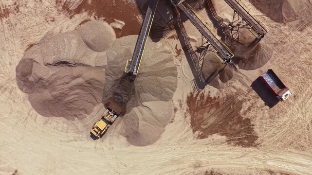 A large truck unloads sand piles at a construction site surrounded by loose dirt. Conveyor belts transport materials across the site. The work occurs in daylight.