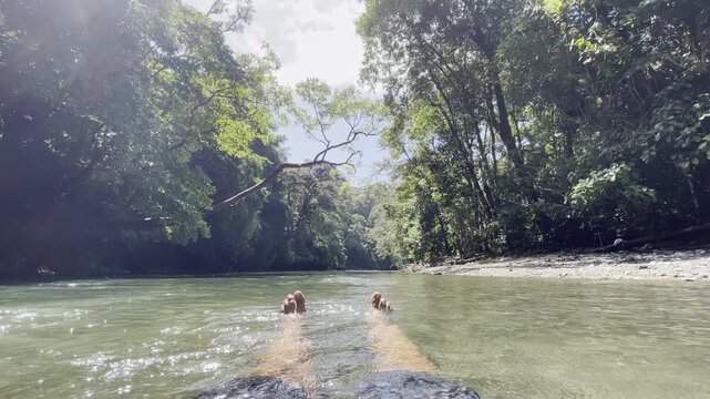 Relaxed POV feet dangle in sparkling jungle river surrounded by dense green rainforest canopy under bright sun creating peaceful Costa Rica nature immersion