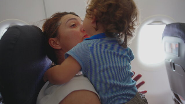 Mother holding restless curly-haired toddler while seated on airplane during daytime flight, trying to comfort fussy child during challenging family travel moment