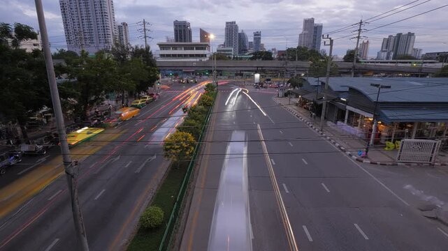 A high-angle overhead time-lapse of Kamphaeng Phet 2 Road near Chatuchak Market intersection at dusk. Rushing traffic transforms into colorful lines of light