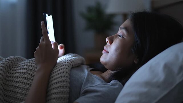 Asian Woman Using Smartphone in Bed at Night, Blue Light from Screen Illuminating Face