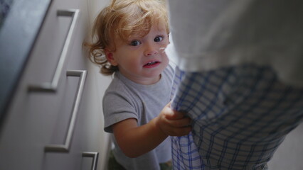 Fototapeta premium Toddler standing near mother in kitchen while she cooks, holding onto her clothes and seeking attention during candid authentic family moment at home