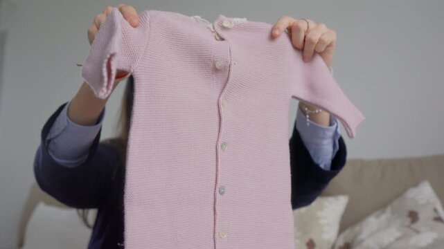 Young woman holding up a soft pink knitted baby romper with button fastening. Newborn clothing preparation and maternity lifestyle concept