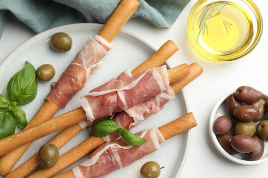 Grissini breadsticks with prosciutto, olives, oil and basil on white table, flat lay