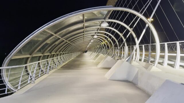 Long perspective view inside illuminated modern arched pedestrian bridge at night with repeating metal structure