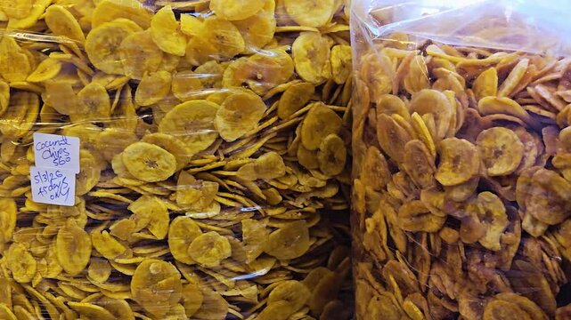 Crispy golden murukku, sweet salted Nendran banana chips, & coconut chips snacks fill clear retail bags at a Kerala market stall. Hand-written price tags show pricing, quantity, & pkg/expiry details.