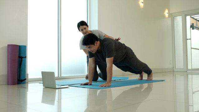 Man performing plank yoga pose on mat at home with partner guidance, strengthening core, arms and shoulders, improving stability and endurance