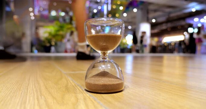 High-energy time-lapse centered on sand timer on shopping mall floor, with sand falling rapidly as blurred shopper silhouettes rush by in background.