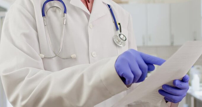 A doctor in a white coat holds paper test results in his hands, examines the tests, and shifts papers in his hands while standing in the office.