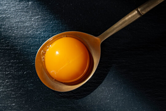 A vibrant yellow egg yolk sits in a vintage metal spoon against a dark slate texture. Dramatic lighting emphasizes freshness and gourmet cooking concepts.