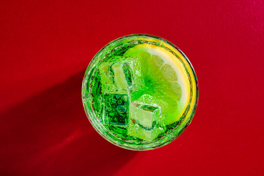 Green soda with lemon slice on red background. Vibrant top view of a bubbly green beverage with a fresh lemon. Ideal for summer drink menus, cocktail advertisements and refreshing beverage concepts