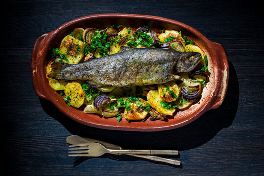Baked whole trout with potatoes and red onion in ceramic dish. Roasted whole rainbow trout served on bed of baked potatoes and onions with fresh herbs. Rustic healthy seafood dinner concept