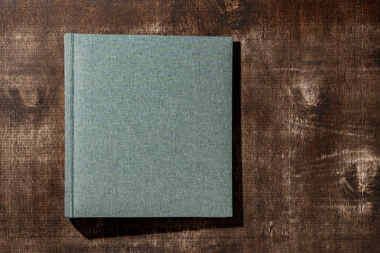Grey green linen book cover on wooden background. Closed fabric photo album or notebook on rustic wooden surface with hard shadows. Minimalist mockup for book design portfolio and memory concepts