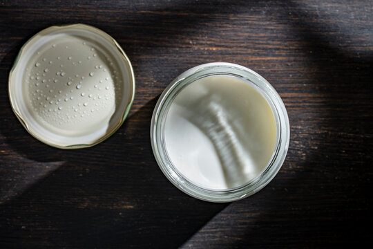 Open glass jar of creamy white yogurt on dark wood. Top view of glass jar with thick white yogurt or sour cream on dark wooden background with shadows and condensation droplets on the lid