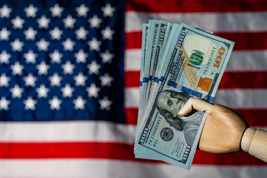 Wooden hand holding hundred dollar bills in front of US flag. Mannequin hand holding a fan of 100 dollar banknotes against blurred American flag. Concept of US economy finance wealth and investment