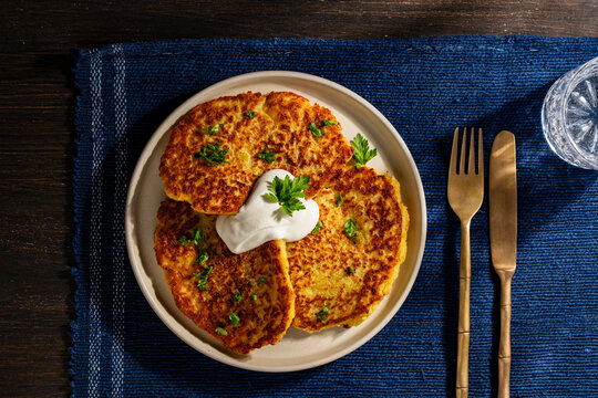 Crispy golden potato pancakes or draniki served with a dollop of sour cream and fresh green herbs on a plate on blue napkin, closeup. Traditional rustic breakfast concept on dark wooden background