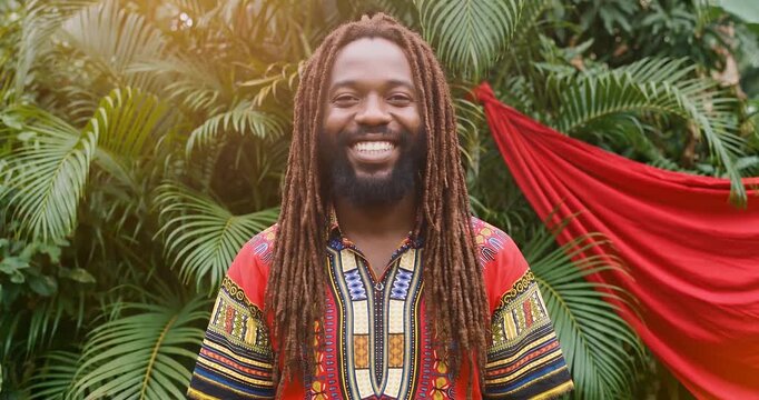 Broad smiling man with long dreadlocks in front of tropical plants expressing genuine happiness and authentic style