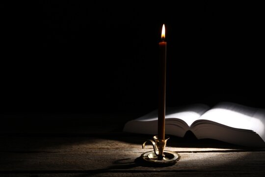 Burning candle and Holy Bible on wooden table in darkness. Space for text