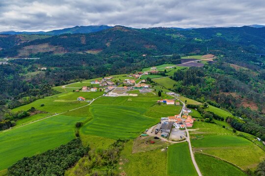 Aerial view of a small rural village nestled among lush green rolling hills and agricultural fields under a cloudy sky in Cudillero, Asturias, Spain.