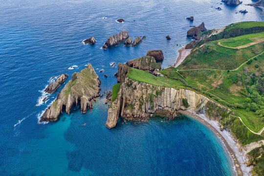 Aerial view of Silence Beach Viewpoint with dramatic vertical cliffs, rocky islets in turquoise water, and lush green hills Cudillero, Asturias, Spain.