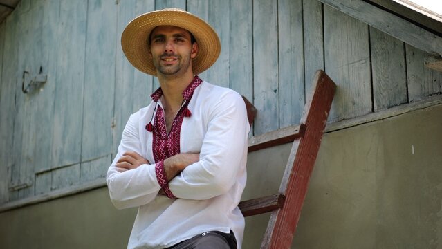 Portrait of smiling guy in straw hat against the background of his house. Young ukrainian man wearing an embroidered shirt looking into camera outdoor. Concept of national identity and patriotism