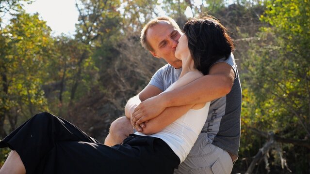 Happy couple hugs during sitting on wooden log on sandy beach. Romantic man kissing his beloved near coastline. Carefree lovers spending time together at resort. Concept of summer vacation or holiday