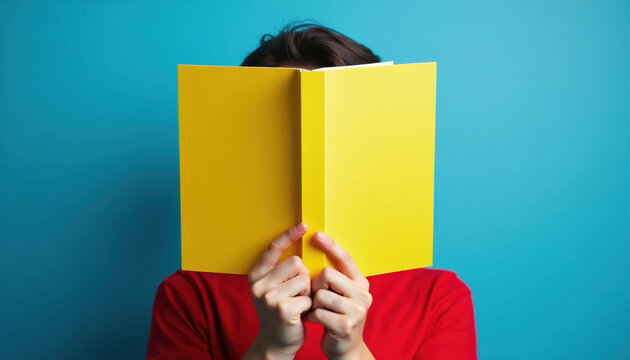 Person holding yellow book covers face. Red shirt contrasts blue background. Mystery reader enjoys literature and learning, hiding identity while absorbed in story.