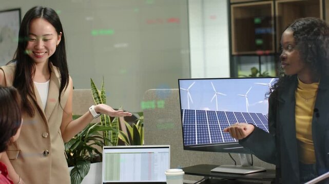 Pair presenting, seated colleague turning and speaking, laptop and solar monitor aiding energy plan