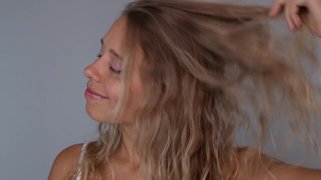 Young blonde woman lifting and tousling wavy hair with both hands, smiling at camera. Playful mood, natural volume and texture, carefree beauty and hair styling concept