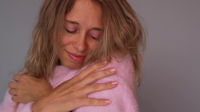 Young blonde woman hugging herself in soft pink sweater, enjoying cozy warmth and gentle touch. Tender mood, self care, comfort and fabric softness concept