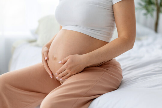 Close-up of pregnant woman sitting on bed tenderly cradling bare round belly with both hands, anonymous cropped image showing third trimester bump in bright home bedroom setting