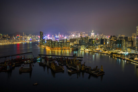 Industrial barges and cranes cluster in Victoria Harbour against the twilight skyline of Hong Kong representing the city maritime and economic strength.