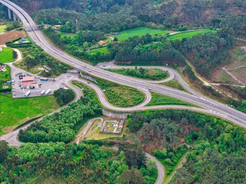 Aerial view of a highway interchange with winding roads and a bridge surrounded by lush green forests and hills in Cudillero, Asturias, Spain.