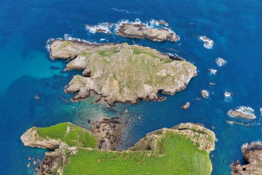 Aerial view of Silence Beach Viewpoint featuring rugged rocky islets and green cliffs surrounded by deep blue ocean water and crashing waves in Cudillero, Asturias, Spain.