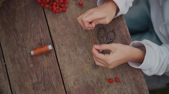Hands threading red rowan berries garland, closeup of artisan creating seasonal decoration with spool of twine on rustic wooden table, cozy autumn mood, slow meditative movements, detailed sleeve