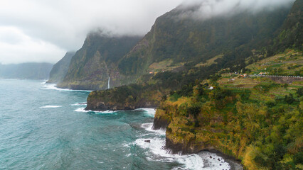 Aerial perspective of the rugged volcanic cliffs on Madeira island featuring a waterfall flowing into the Atlantic Ocean under a heavy overcast sky in Portugal. © JULIA