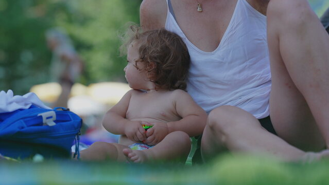 Curious toddler sitting on grass with toy while mother nearby, focused expression during summer picnic, playful childhood moment filled with exploration and discovery