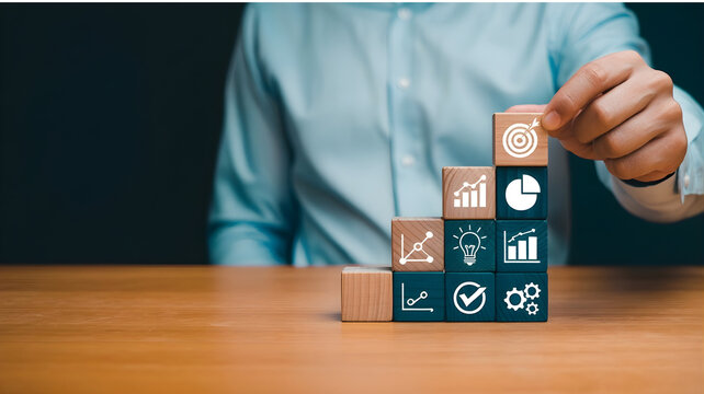 A man in a blue shirt places a block on a stack of blocks with business icons on a table indoors