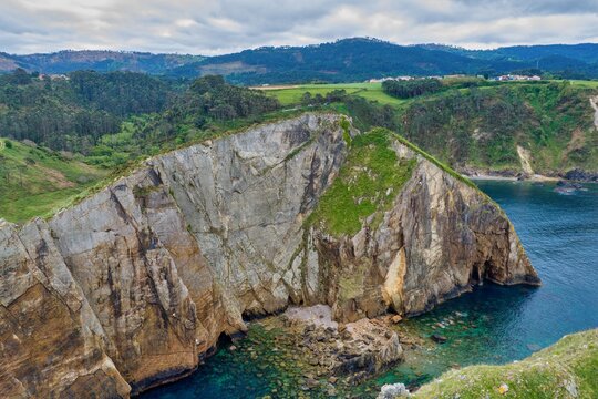 Aerial view of Silence Beach Viewpoint featuring dramatic jagged cliffs, turquoise ocean water, and lush green hills under a cloudy sky in Cudillero, Asturias, Spain.