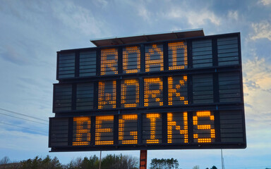 Electronic road sign displaying ROAD WORK BEGINS in bright orange text against a blue sky. Traffic control, infrastructure, construction, and safety themes. Design Element. © mailcaroline