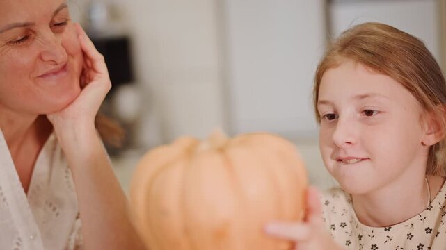 Closeup kitchen caucasian mother daughter pumpkin, softfocus portrait of girl holding small pale pumpkin on wooden table while mother watches with fond smile, warm indoor lighting, seasonal harvest