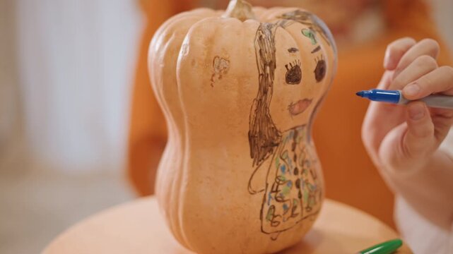 Hands drawing face on squash pumpkin, closeup of textured gourd on wooden table, blue marker creating simple portrait, cozy indoor autumn craft, warm lighting, seasonal decoration idea for home