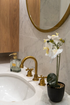 Elegant bathroom vanity scene featuring a gold faucet and round mirror over a marble sink, accented by a white orchid and wooden cabinet for a warm, modern and luxurious feel.
